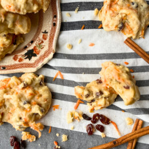 Some carrot and raisin cookies on a grey and white backdrop. There are some cookies on a plate and one is split in half.