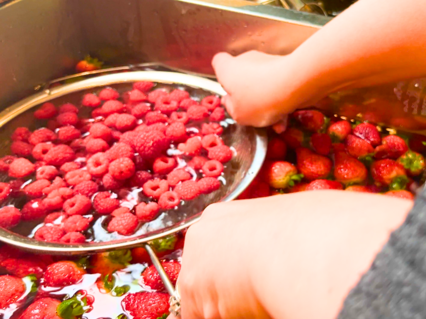 A woman immersing a colander full of raspberries into a sink full of water and strawberries.