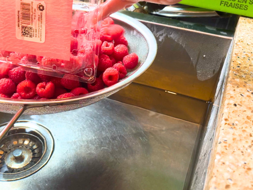 A woman pouring raspberries into a colander overtop of a sink with water.