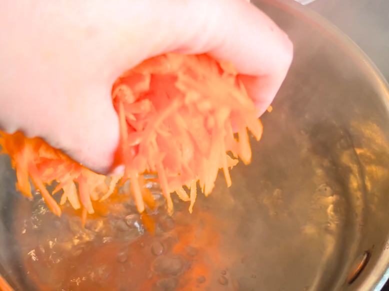 A woman adding shredded carrots to boiling water.