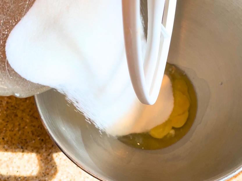Pouring sugar in to a mixing bowl with eggs in the bottom.