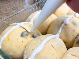 A woman using a piping bag to draw a white cross on bun dough.