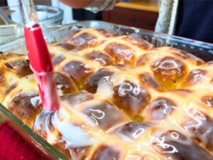 A woman painting a sugar glaze onto a casserole dish of hot cross buns. She is using a red pastry brush.