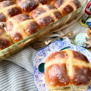 A casserole dish of hot cross buns in the back ground. There is one bun on a blue floral plate.