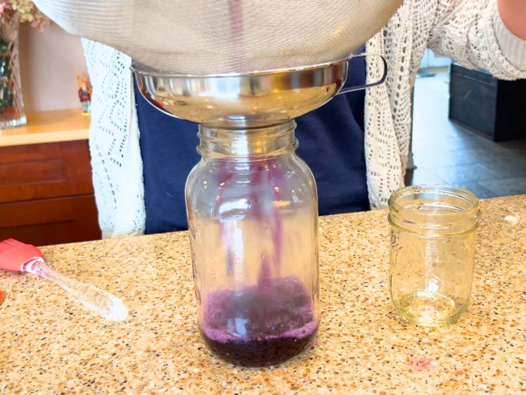 A woman pouring purple liquid through a strainer into a canning jar.