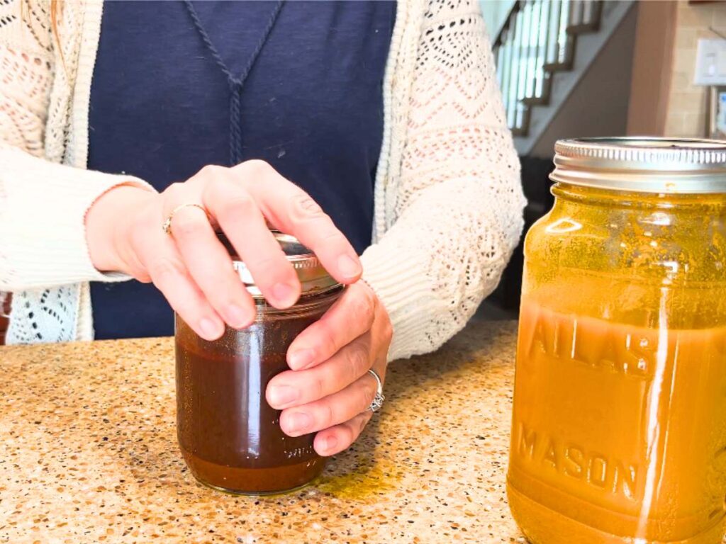 A woman screwing on a lid to a canning jar with brownish-red liquid inside. There is a jar off to the side with yellow liquid inside.