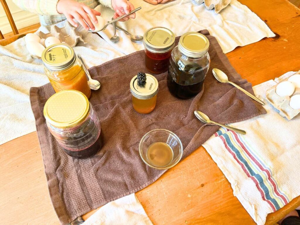 A wooden table with kitchen towels spread out on it. There are 5 canning jars with different coloured liquid inside. There is a girl balancing an egg on a spoon.