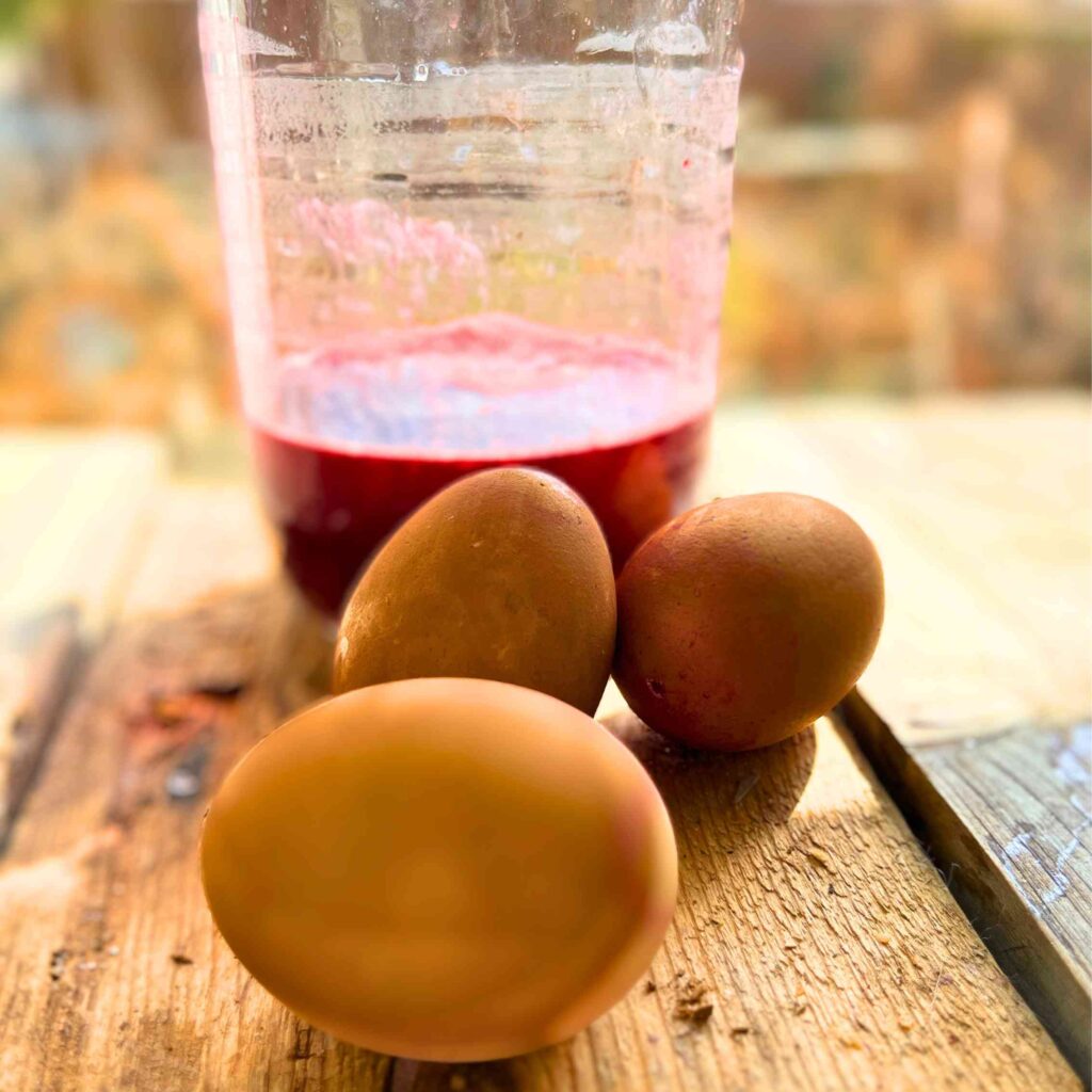 A jam jar with red coloured liquid inside. There are three coloured eggs in front.