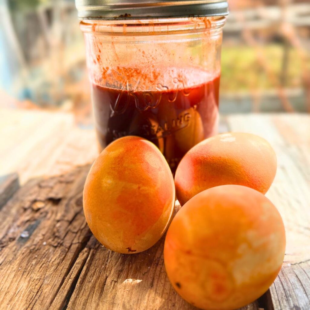 A mason jar with brownish- red liquid inside. There are three orange eggs in front.