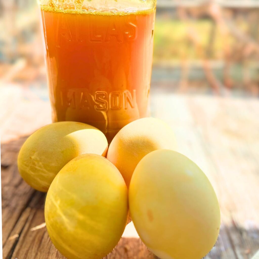 A Mason jar with yellow liquid inside. There are four yellow eggs in the foreground.