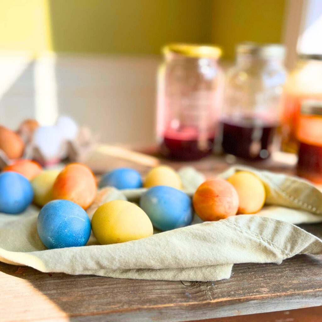 A collection of coloured eggs on a grey cloth, there are some jam jars in the background with coloured liquid inside.
