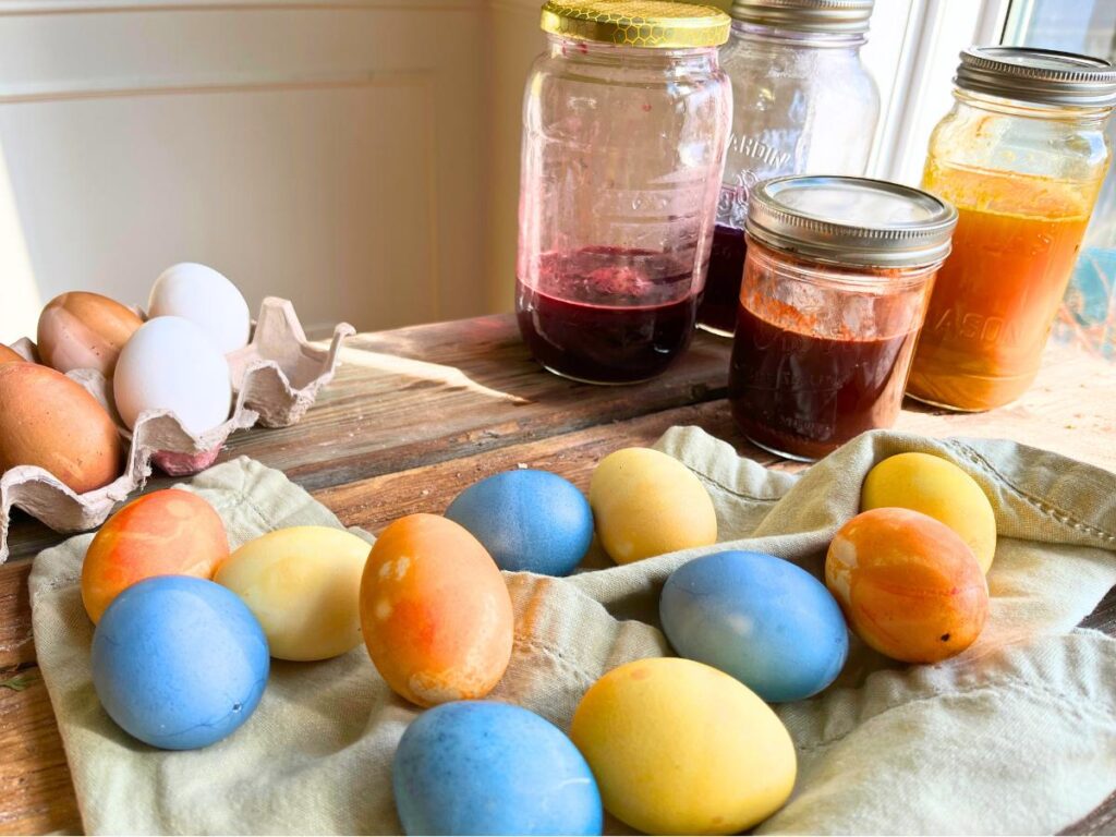 A wooden table with a collection of coloured eggs on top. There are canning jars with coloured liquid in the background.