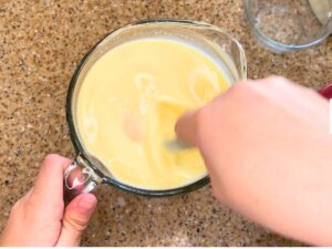 Woman stirring a light yellow liquid inside of a glass measuring cup.