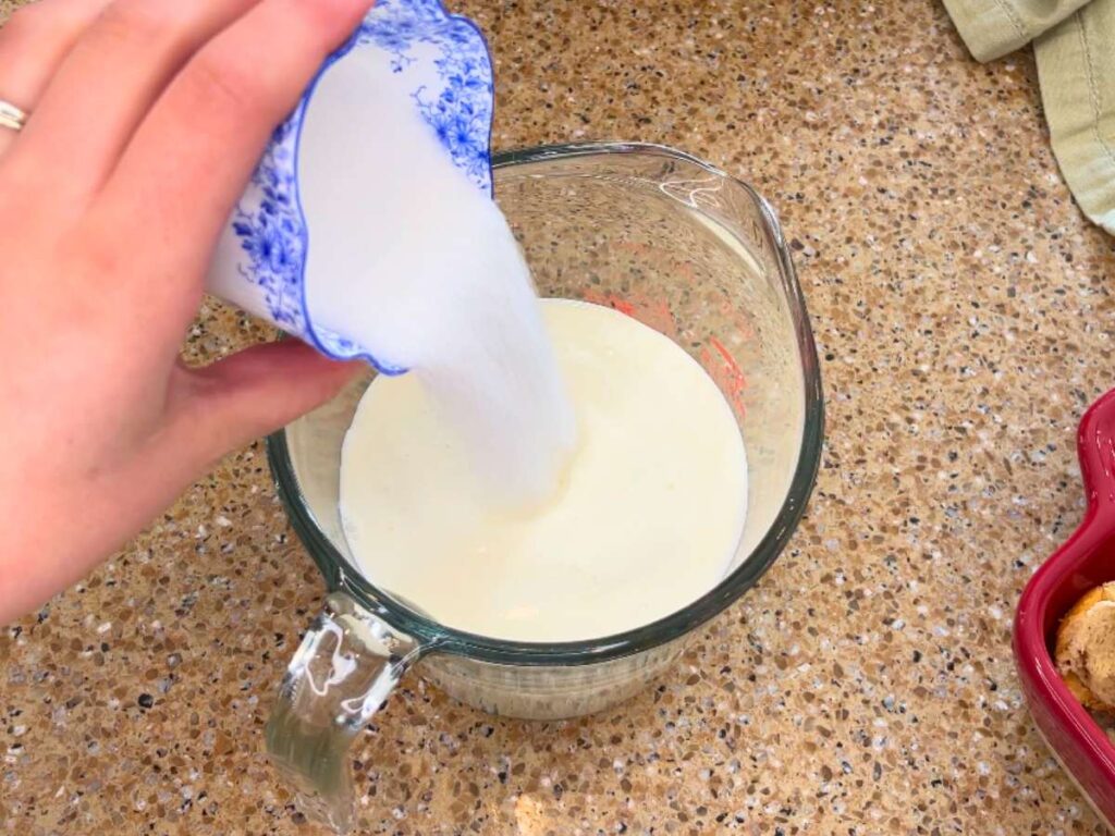 Woman adding a teacup with sugar to a glass measuring cup with milk inside.