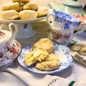 Some biscuits on a platter. There are tea cups and a tea pot in the background. There is a plate in the foreground with some split biscuits on it with butter.