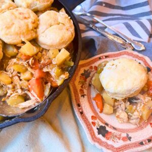 A cast iron skillet with a chicken and vegetable mixture inside. There are biscuits on the top. There is a plate in the foreground with a scoop of the mixture and a biscuit on top.