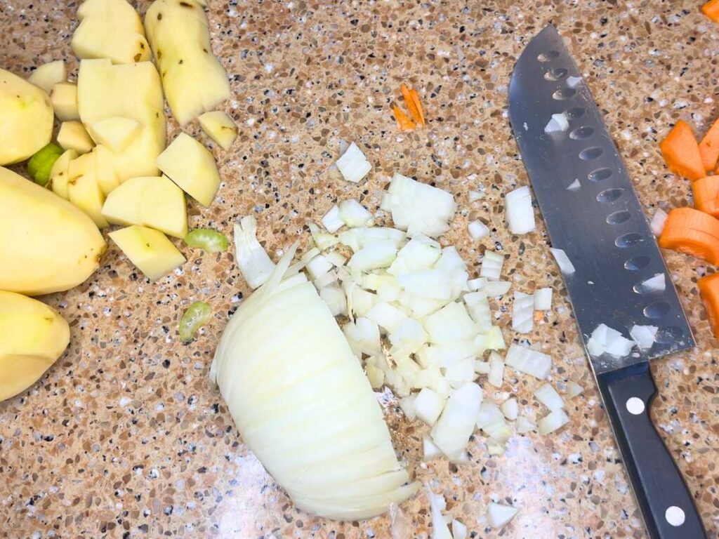 A half chopped white onion on a counter top. The knife is beside the onion. There are chopped potatoes and carrots to the side.