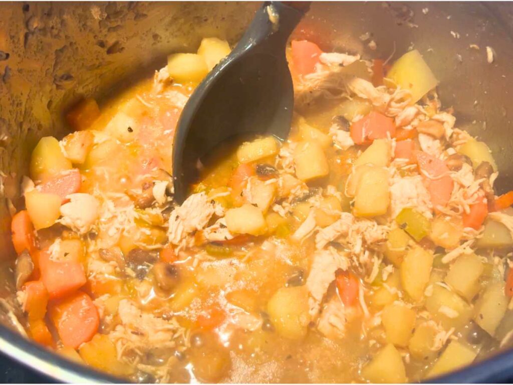 A creamy saucy mixture of chicken and vegetables in a large metal pot being stirred by a large grey spoon.