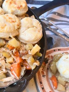 A skillet with a cooked chicken and vegetable mixture inside. There are baked biscuits on top. Beside in a brown floral plate with a scoop of the mixture and a biscuit on top.