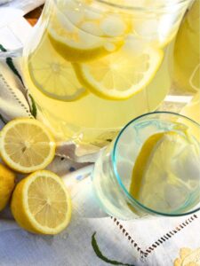 A pitcher of lemonade with lemon slices inside. In the foreground there is a glass with ice, a lemon slice and liquid inside.
