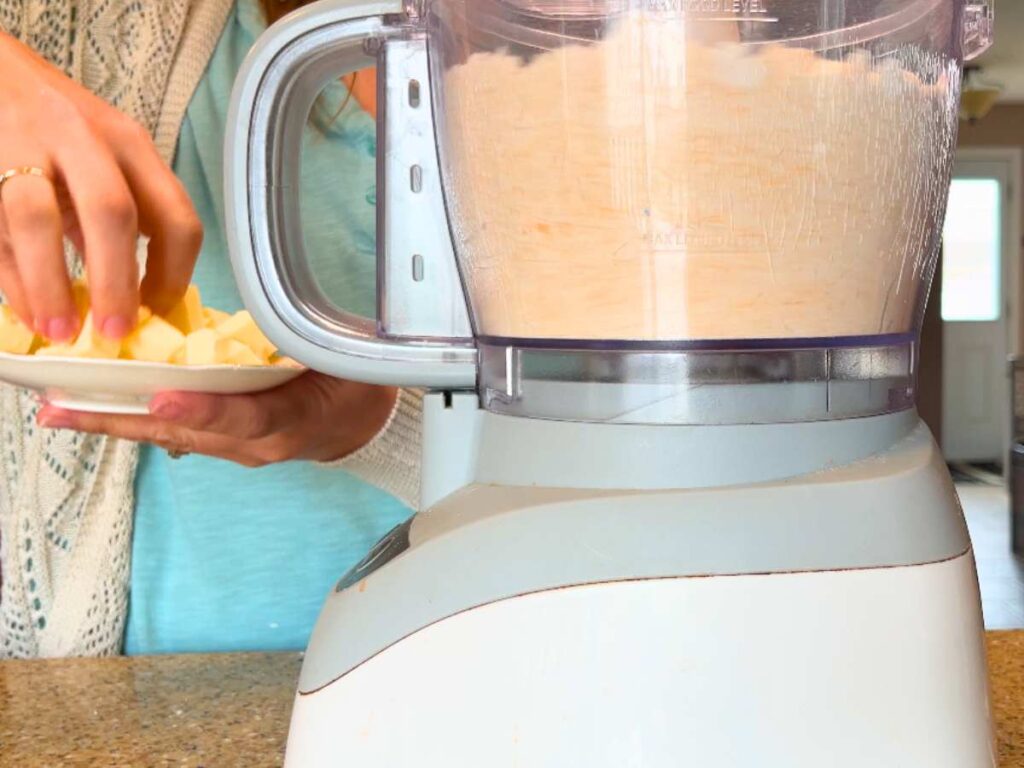 A woman adding cubes of butter to a food processor full of a dry mixture.
