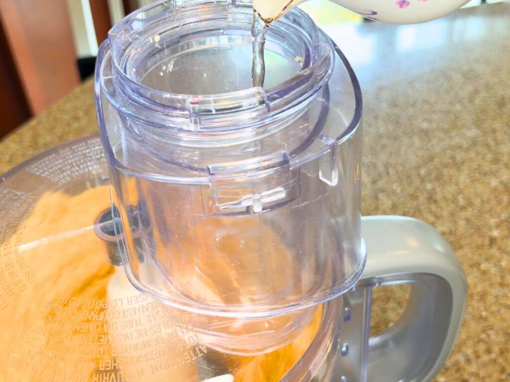 A woman pouring a small amount of water into a food processor.