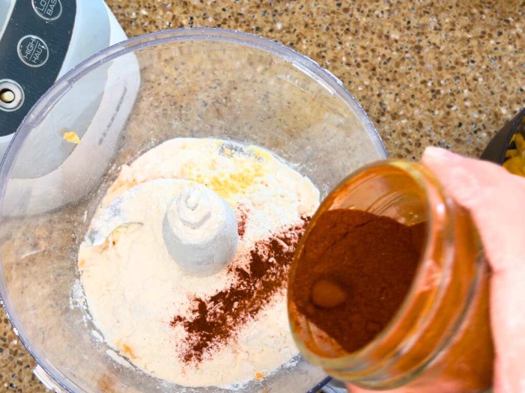 A woman adding a reddish-brown powder to a food processor with flour in it.