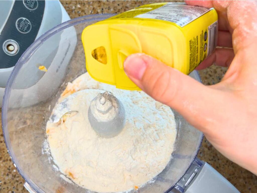 A woman adding a powder on a yellow tin to a food processor with flour in it