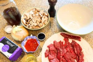 A countertop full of ingredients including raw beef strips, oil, tomato sauce, and onion, butter, sliced mushrooms, pepper, salt, and flour.