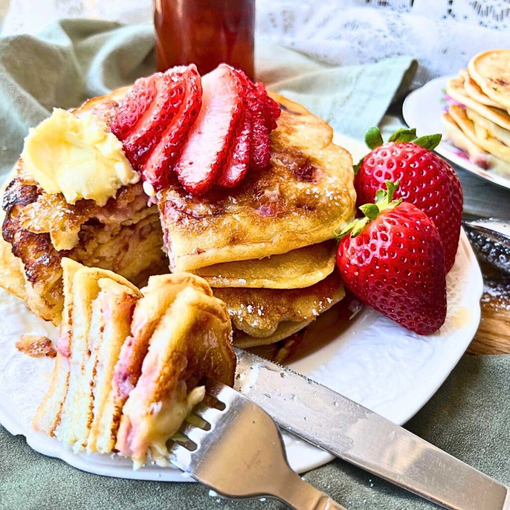 A stack of pancakes on a white plate. There are strawberries on the plate. There is a fork with some of the pancakes on it.