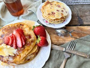 A stack of pancakes on a white plate. There are strawberries on the plate. There is a smaller plate of pancakes behind.