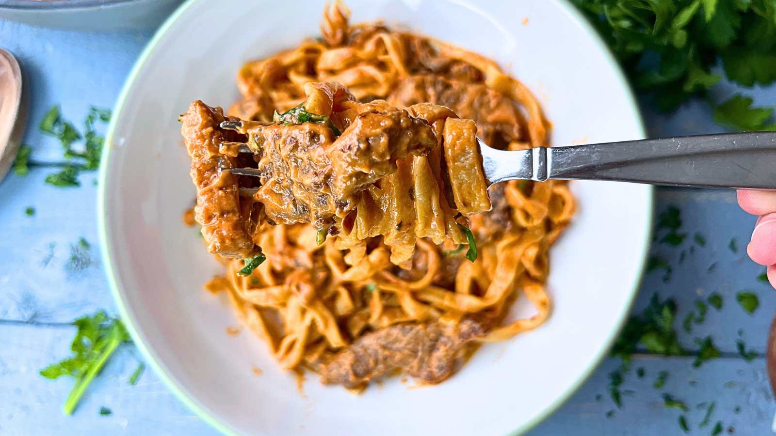 A large white bowl with a beef stroganoff pasta inside. There is a woman holding up a forkful of pasta and beef.