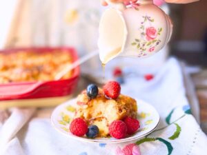 A woman pouring syrup from a small floral pitcher. There is a red casserole in the background. A portion of it is on a yellow floral plate with berries and syrup.