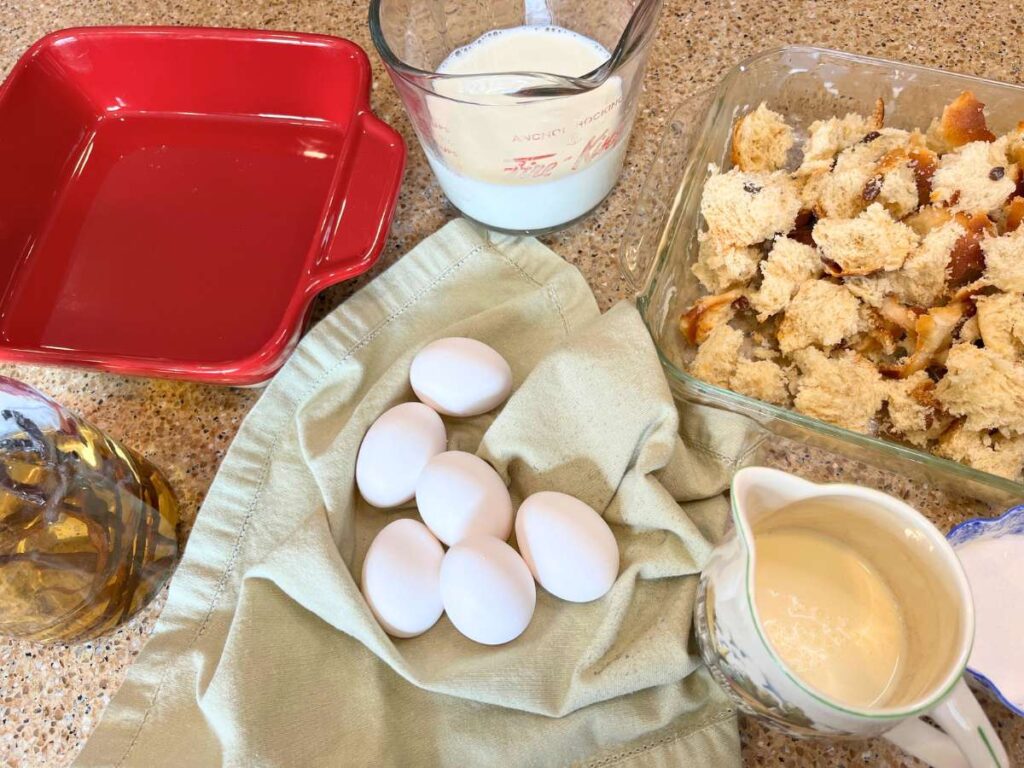 A counter top with a red casserole dish, vanilla, eggs, milk, cream, sugar, and chunks of bread.