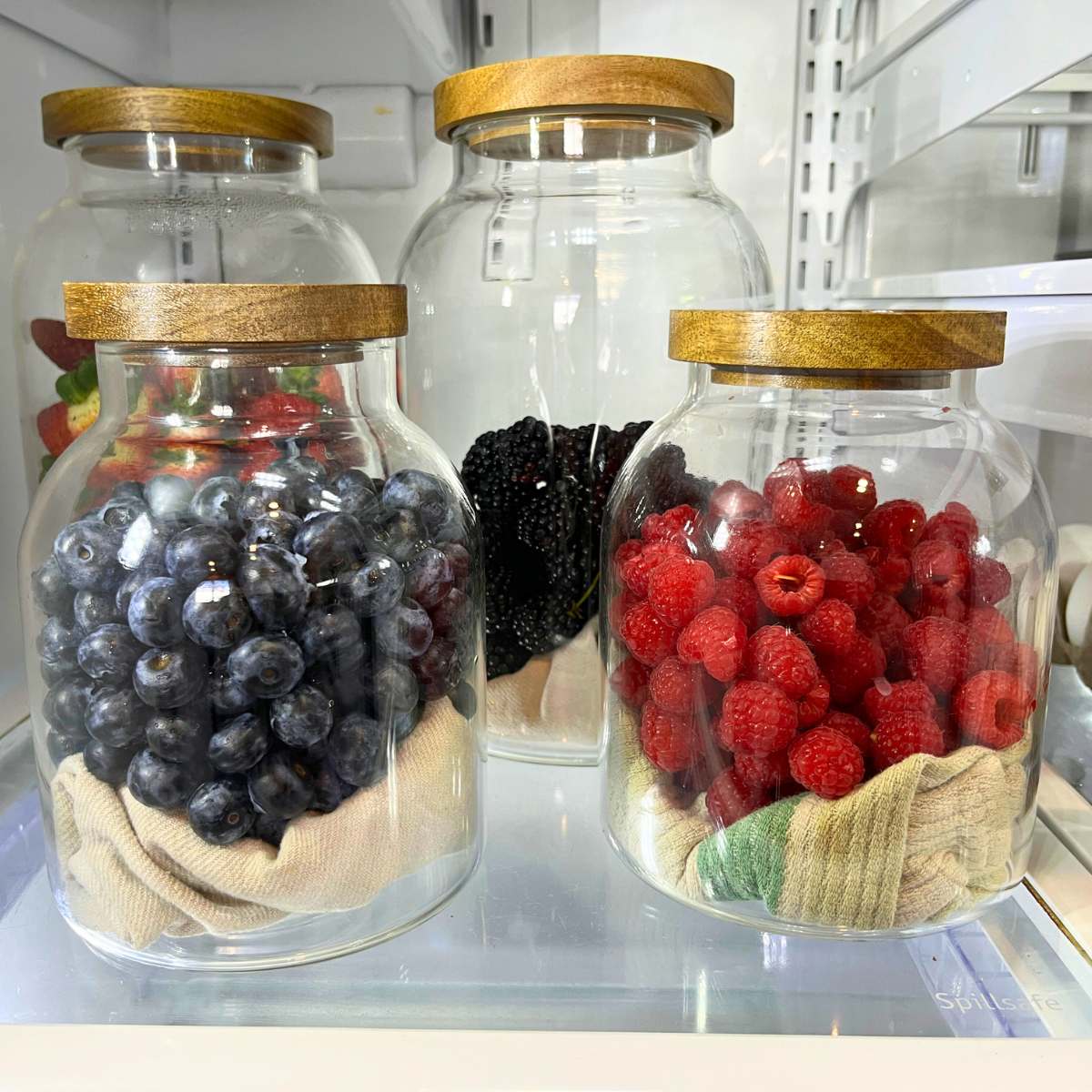Four glass jars with wooden lids full of berries. They all have a kitchen towel in the bottom.