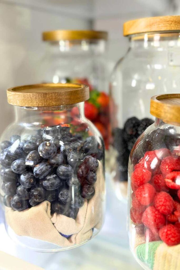 Four glass jars with berries in them. They all have wooden lids and have a kitchen towel in the bottom.