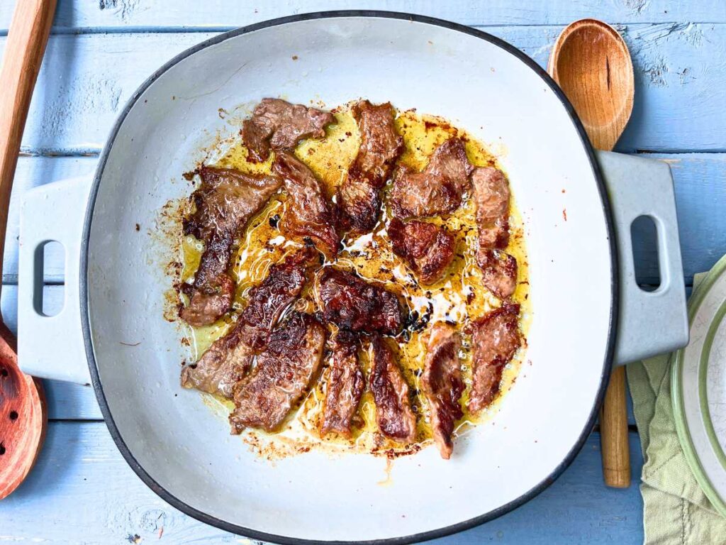 A grey Dutch oven with beef strips frying inside.