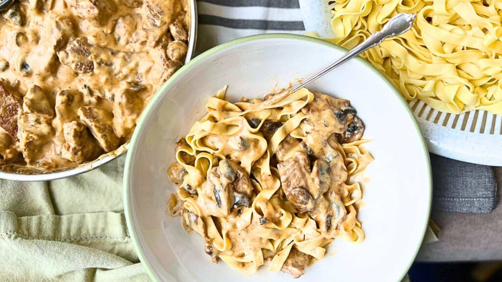 A large white bowl with beef stroganoff pasta inside. There is more stroganoff in a pan to the side and more noodles in a colander to the other side.