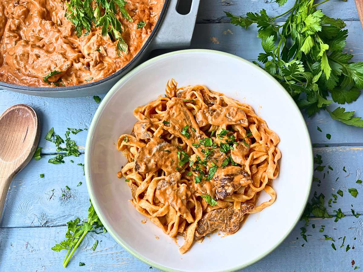 A large white bowl of beef stroganoff mixed with pasta. There is more of the stroganoff to the side in a large sauce pot.