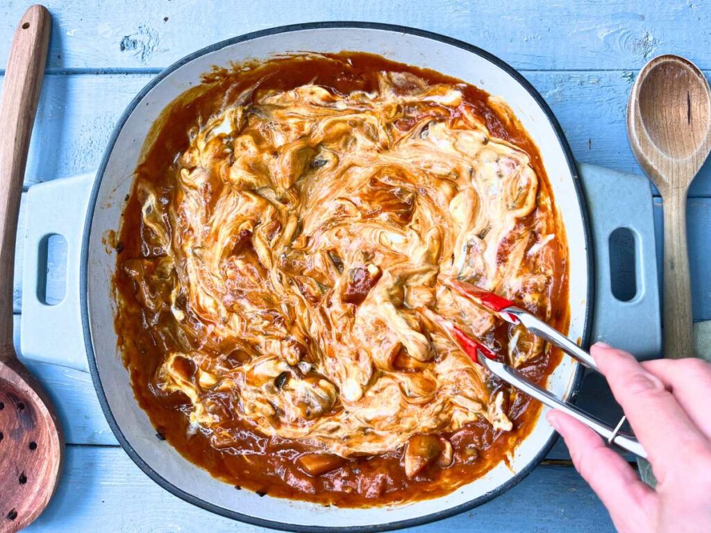 A woman is using tongs to stir sour cream into beef stroganoff.