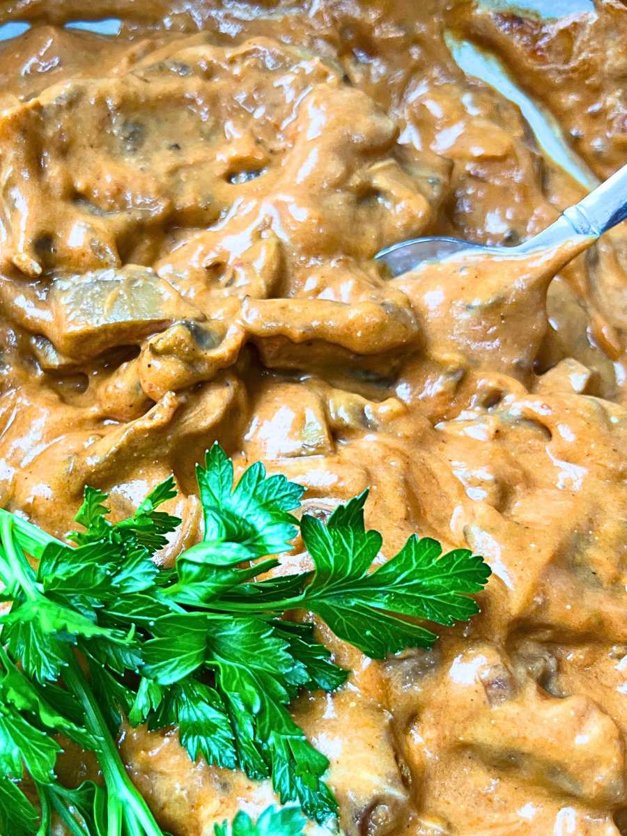 A close up look at a Dutch oven full of cooked beef stroganoff. There is a spoon inside and parsley on top.