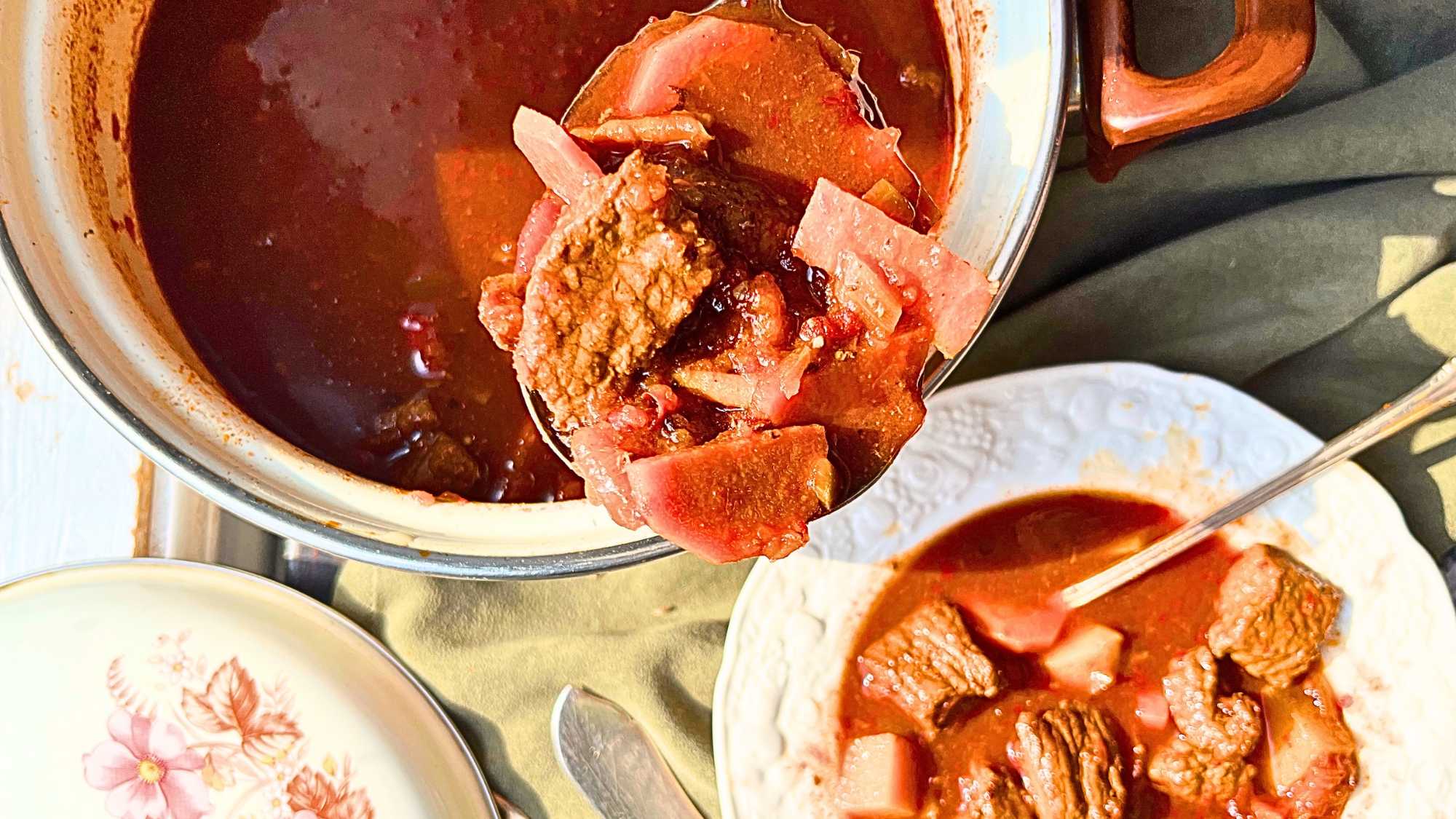 A woman is scooping out some beef goulash. There is a bowl full in the front.