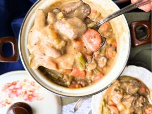 A white floral pot with vegetable beef stew inside. There is a white bowl with a small amount of beef stew.