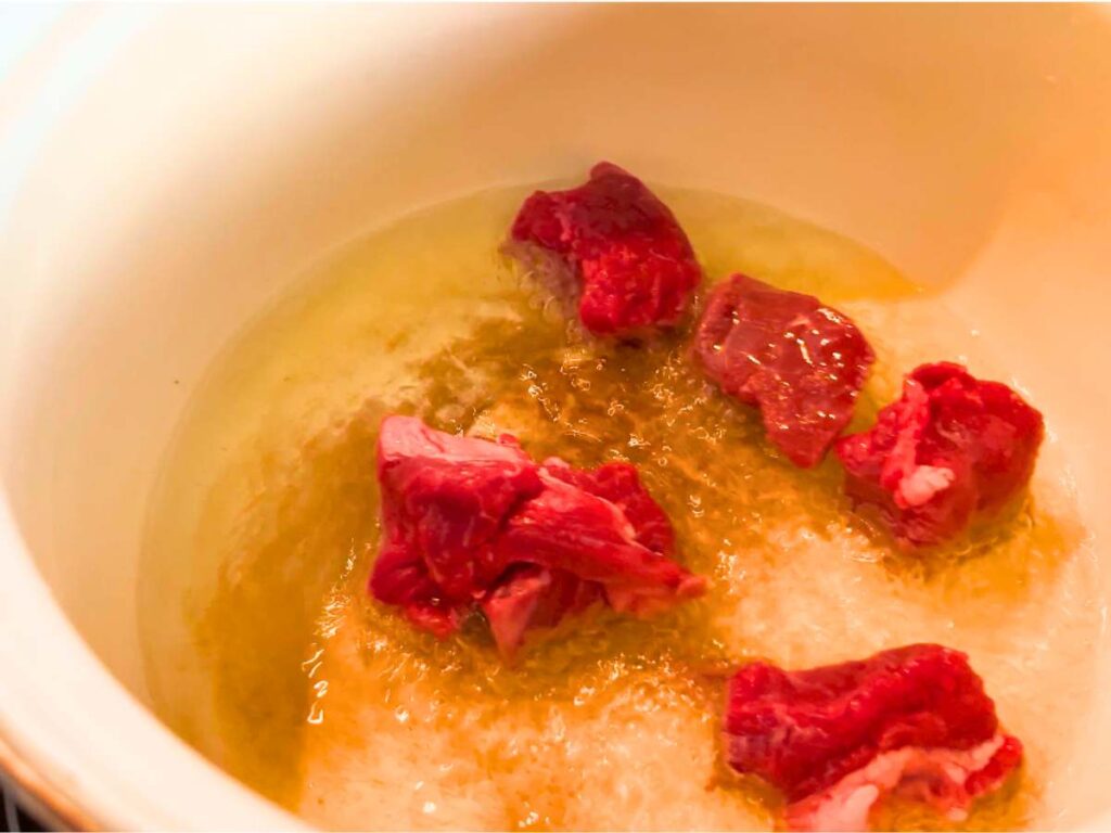 Chunks of raw beef cooking in oil in a white pot.