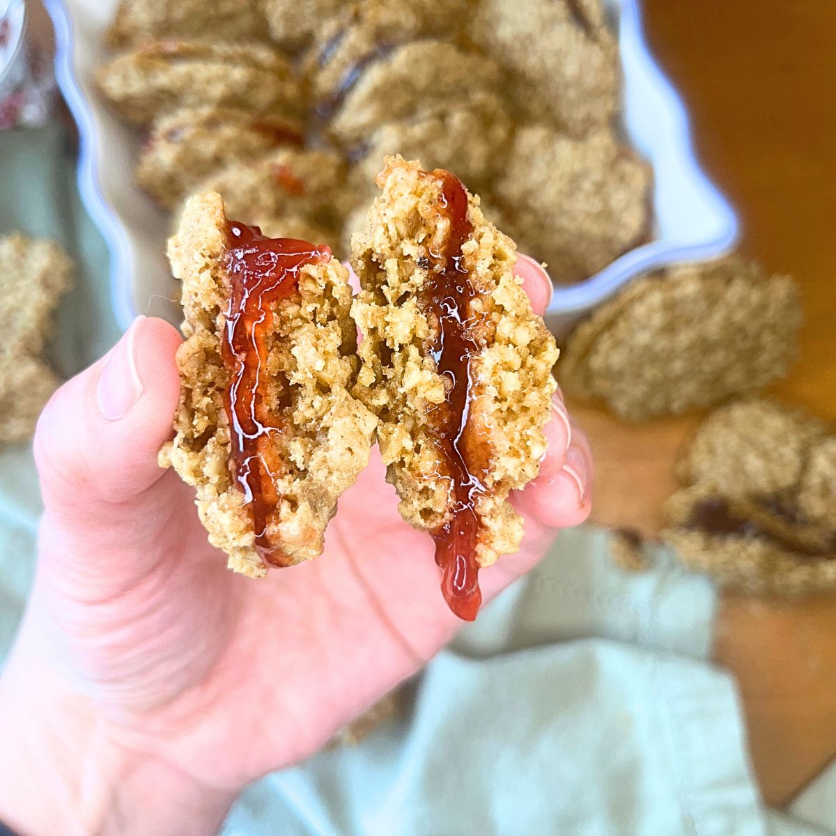 There is a woman holding a split oatmeal cookie sandwiched with jam. There are more cookies in the background.