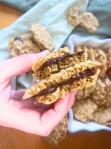 There is a woman holding a oatmeal cookie sandwiched with chocolate. There are more cookies in the background.