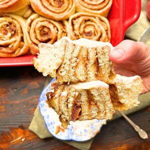 There is a woman holding up a cinnamon roll that has been cut in half. There is white icing on top. There are more cinnamon rolls in a red casserole dish in the background.