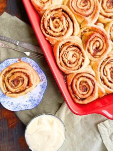 There is a red casserole dish full of cinnamon rolls there is one cinnamon bun an a blue floral plate. There is a small dish full of white icing.