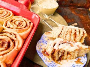 There is a red casserole dish with cinnamon buns inside. On the side there is a blue floral plate with a cinnamon roll that has been cut in half. There is white icing on top.