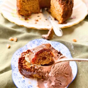 A Bundt cake in the background. In the front there is a slice of cake on a blue floral cake. There is a scoop of chocolate ice cream on top.